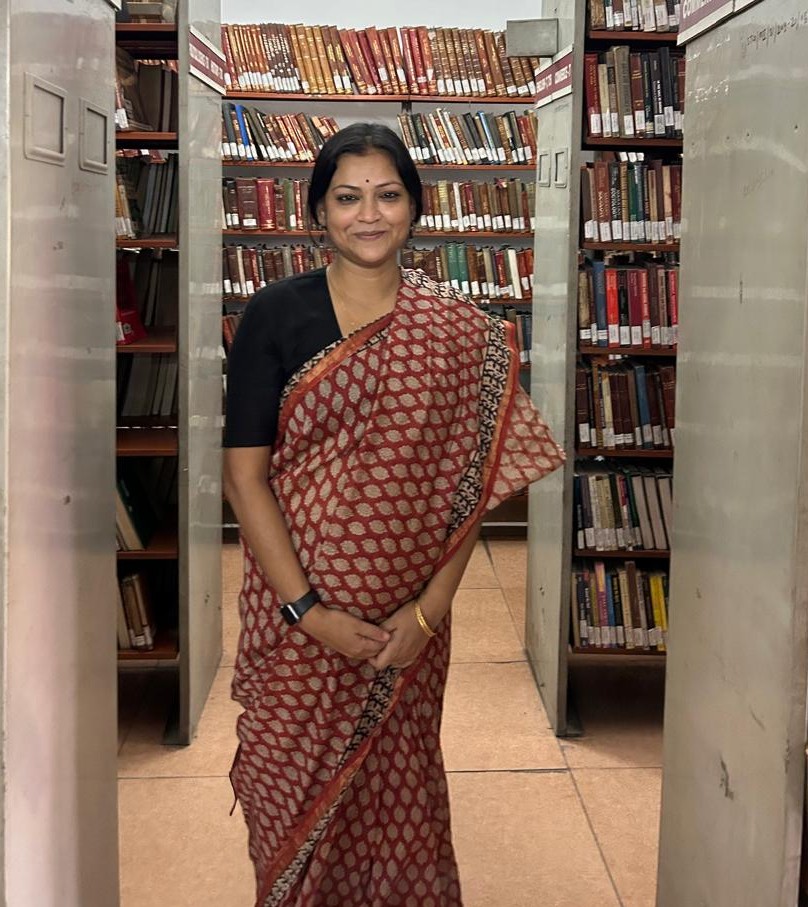 Dr. Priyanka Jha standing in a library aisle between tall bookshelves, wearing a red and beige saree, smiling with hands clasped in front
