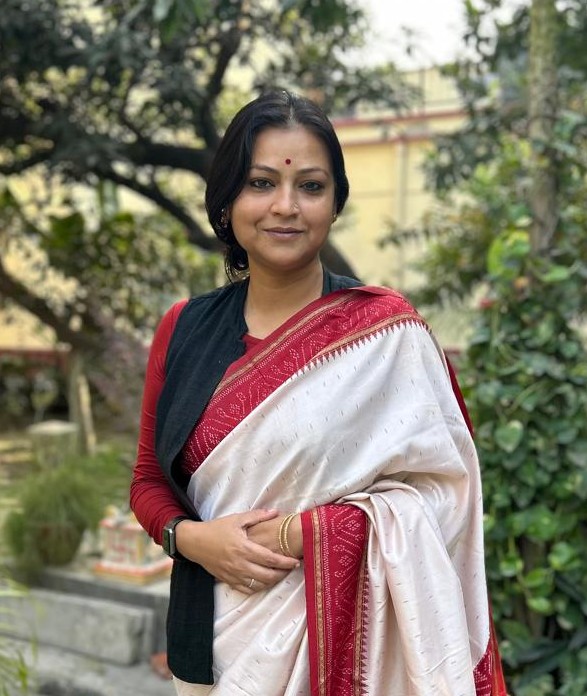 Dr. Priyanka Jha standing outdoors in a garden courtyard, wearing a white saree with red and gold borders, paired with a red blouse and black vest